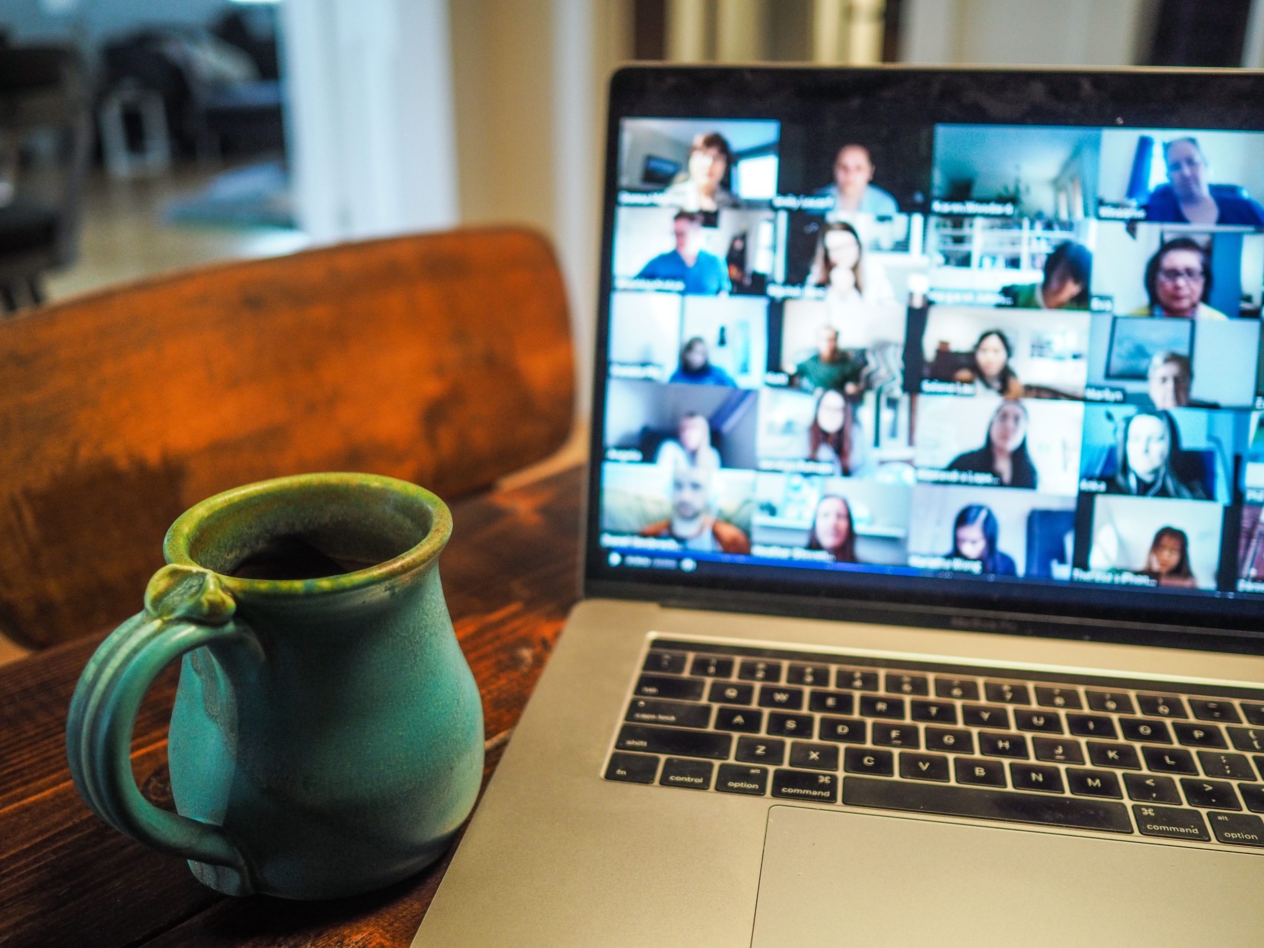 Laptop shows a screen with numerous faces of people all together on a virtual call with a coffee cup in the foreground